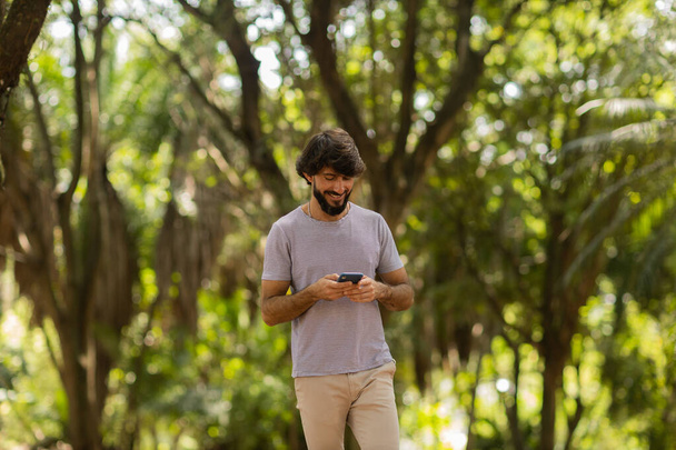 Vue d'un jeune homme utilisant un smartphone le jour avec un parc verdoyant en arrière-plan. Téléphone portable, technologie, concept urbain. Photo de haute qualité - Photo, image
