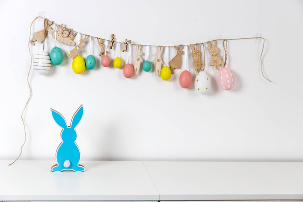 Fragment of the interior. Decorated children's room for Easter. A garland of plastic eggs and hares cut out of cardboard on the wall. Wooden blue rabbits . Place for your text. Easter card. - Foto, Imagen