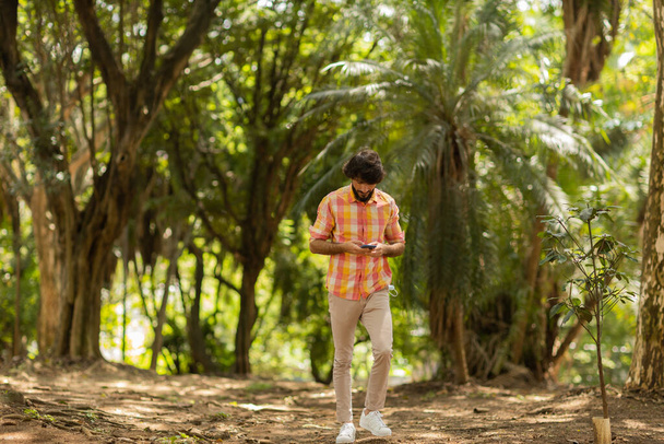 Pohled na mladého muže pomocí smartphonu ve dne se zeleným parkem v pozadí. Mobilní telefon, technologie, městský koncept. Kvalitní fotografie - Fotografie, Obrázek