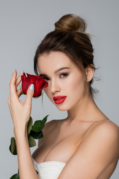 charming woman with bare shoulders and makeup holding red rose near face isolated on grey - Fotó, kép