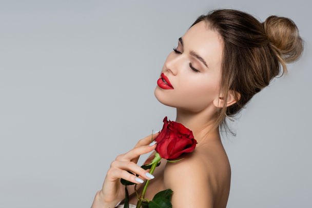 young and pretty woman with closed eyes holding red rose near bare shoulder isolated on grey - Photo, Image