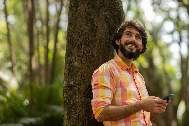Vista de un joven usando un teléfono inteligente durante el día con un parque verde en el fondo. Teléfono móvil, tecnología, concepto urbano. Foto de alta calidad - Foto, imagen