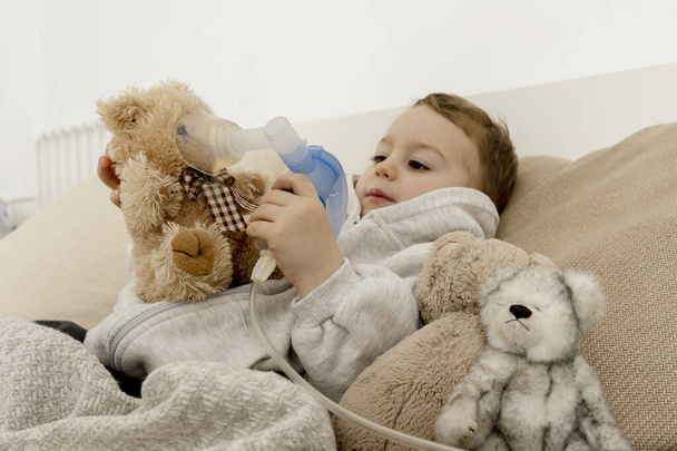 Küçük hasta çocuk öksürük tedavisi için astım spreyi kullanıyor. Hasta çocuk nefes alıp veriyor ve oyuncak ayısını da soluyor. Grip mevsimi. Evde tıbbi prosedür. İç mekan ve giysiler doğal toprak renklerinde.. - Fotoğraf, Görsel