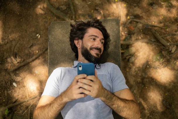 Widok młodego człowieka używającego smartfona w ciągu dnia leżącego na ławce w parku. Wysokiej jakości zdjęcie. Telefon komórkowy, technologia, urbanistyka, koncepcja natury. Wysokiej jakości zdjęcie - Zdjęcie, obraz