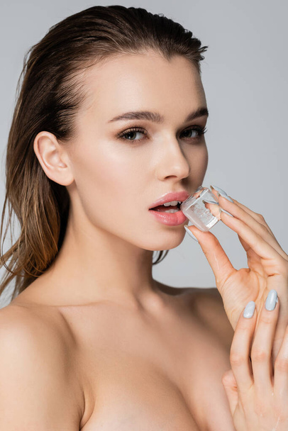 charming woman with wet hair holding ice cube near lips while looking at camera isolated on grey - Photo, Image