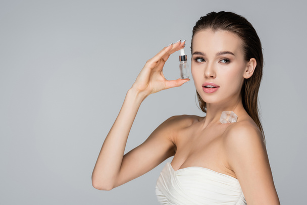 young woman with cosmetic serum and ice cube on bare shoulder looking away isolated on grey - Фото, изображение