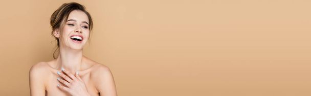 mujer alegre con hombros desnudos sosteniendo la mano en el pecho y riendo aislado en beige, pancarta - Foto, Imagen