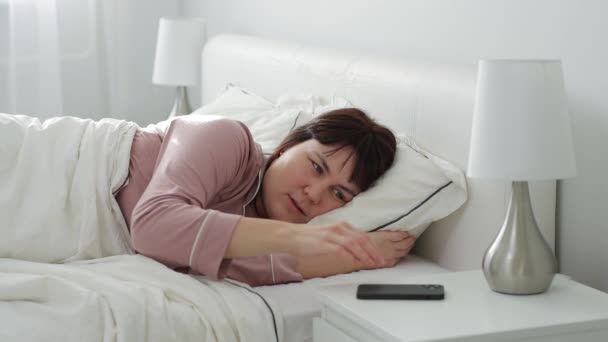 Концепция доброго утра - молодая женщина просыпается, чтобы выключить будильник на своем смартфоне и ложится спать  - Кадры, видео