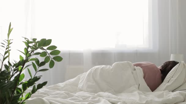 buen concepto de la mañana - mujer joven en pijama rosa despertando y estirándose en dormitorio luminoso - Metraje, vídeo