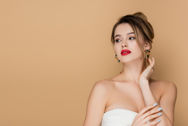 pretty young woman in strapless top and golden earrings looking away isolated on beige - Photo, Image