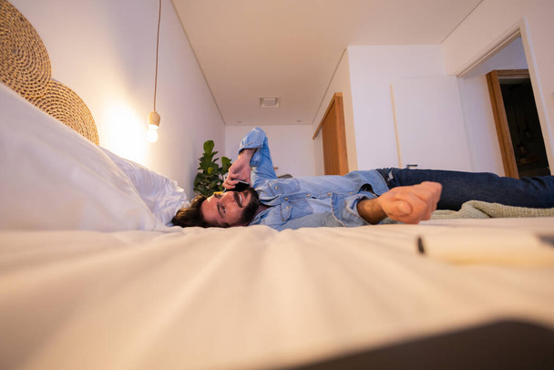 Widok z boku młodego człowieka korzystającego ze smartfona w nocy ze swoim łóżkiem w sypialni. Telefon komórkowy, technologia, koncepcja. Wysokiej jakości zdjęcie - Zdjęcie, obraz