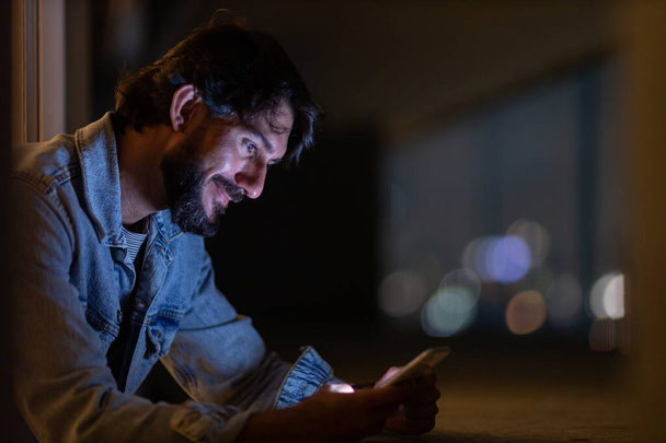 Boční pohled na mladého muže pomocí smartphonu v noci na jeho balkóně s výhledem na město krajinu v pozadí. Mobilní telefon, technologie, koncept. Kvalitní fotografie - Fotografie, Obrázek