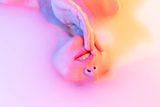 Closeup female face, young beautiful girl in milk bath with soft glow in blue-green neon light. Beauty, fashion, style, skincare concept - Photo, Image