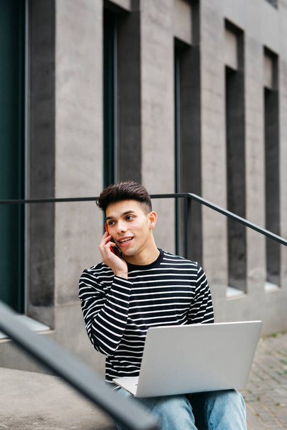 Молодой человек, сидит на улице, пользуется ноутбуком и разговаривает на красном смартфоне - Фото, изображение