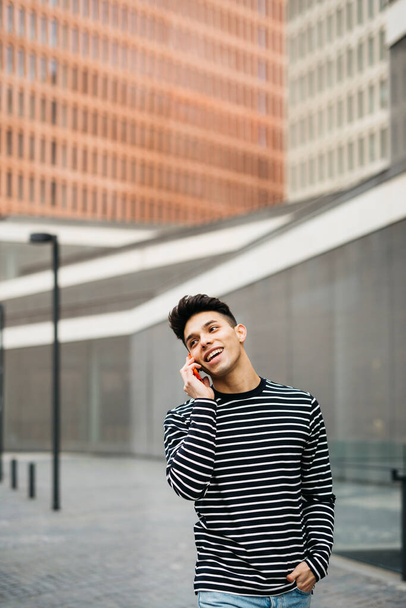 Jonge glimlachende man lopen en praten op een rode smartphone de een straat - Foto, afbeelding