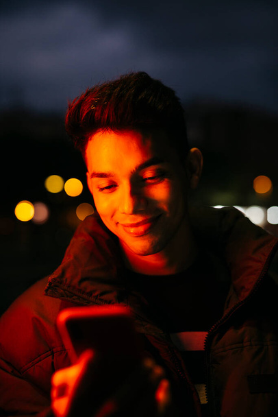 Портрет молодого человека на улице в черном пальто и с помощью смартфона, под красным светом - Фото, изображение