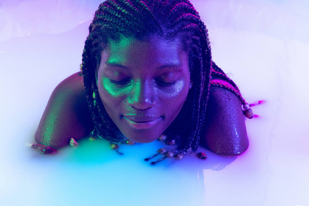One beautiful young dark skinned girl in milk bath with soft glow in blue-white neon light. Copyspace for advertising. Beauty, fashion, style, skincare concept - Фото, зображення