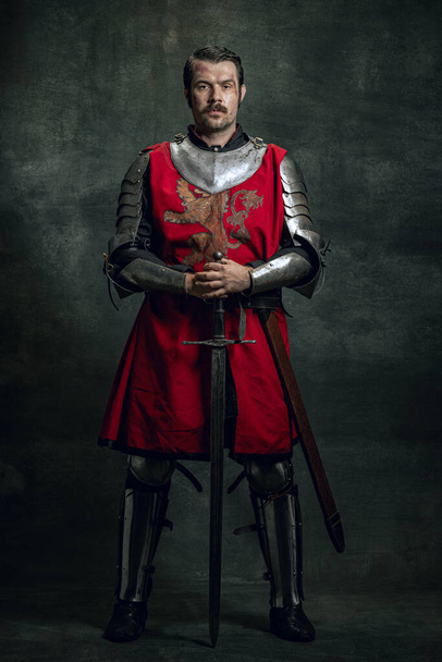 Portrait of brutal seriuos man, medieval warrior or knight with dirty wounded face holding sword isolated over dark background. Comparison of eras - Fotografie, Obrázek