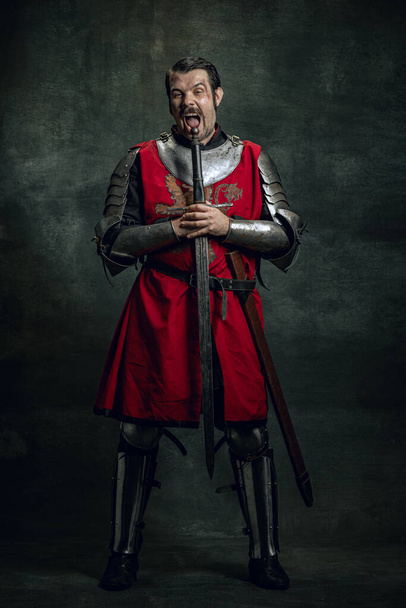 Comic portrait of funny medieval warrior or knight with dirty wounded face holding sword isolated over dark background. Comparison of eras - Fotografie, Obrázek
