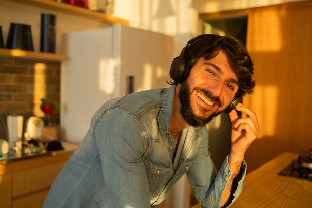 View of young man listening to music with headphones in his ears in the kitchen of his apartment. High quality photo. Mobile phone, technology, music, urban living, feel good concept. - Foto, afbeelding