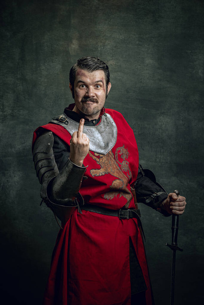 Comic portrait of funny medieval warrior or knight with dirty wounded face gesturing isolated over dark background. Comparison of eras - Foto, imagen