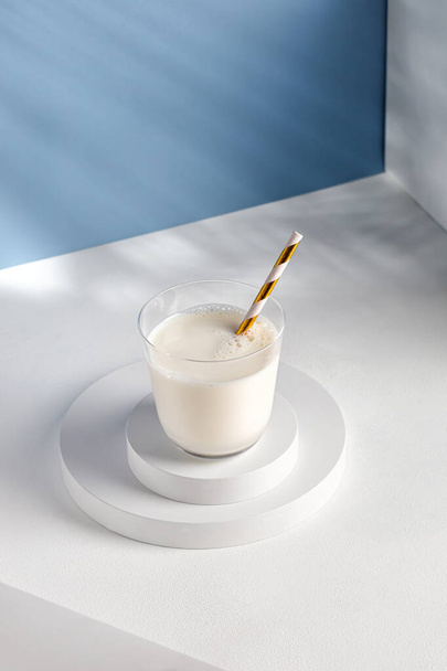 Milk in glass with a straw on white podiums. Natural milk on pedestal in sunlight. Shadow on blue wall. - Фото, изображение