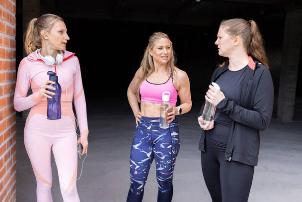 Sporty beautiful young women of different body types talking and drinking water from bottle after working out outside of a gym, gym, fitness, sport concept  - Photo, Image
