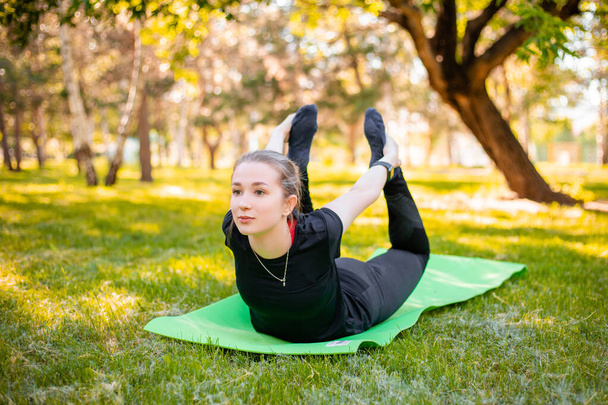 Outdoor fitness and pilates workout. A young woman does exercises to stretch the muscle,  while sitting on a yoga mat in the park. Exercise to develop flexibility.  - Foto, Imagen