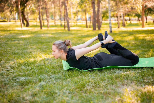 Outdoor fitness and pilates workout. A young woman does exercises to stretch the muscle,  while sitting on a yoga mat in the park. Exercise to develop flexibility.  - Fotoğraf, Görsel