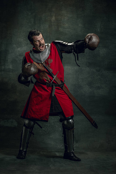 Portrait of medieval warrior or knight with dirty wounded face in boxing gloves isolated over dark background. Comparison of eras, history - Photo, Image