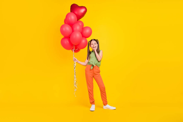 Photo of excited shocked girl dressed green top holding red bubbles bunch hand arm cheek cheekbone isolated yellow color background - Фото, изображение