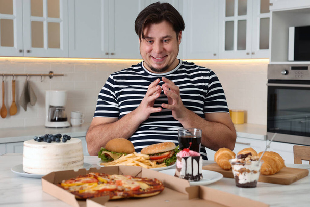 Hungry overweight man at table with sweets and fast food in kitchen - Фото, изображение