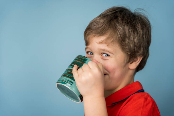 A Caucasian boy in a red T-shirt drinks tea from a mug and smiles fervently looking at the camera. Blue background. - Foto, Imagem