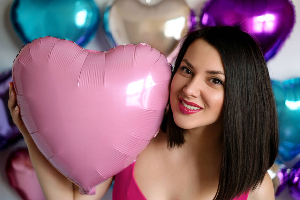 Μια νεαρή όμορφη μελαχρινή γυναίκα κρατά ένα ροζ μπαλόνι σε σχήμα καρδιάς στο χέρι της, χαμογελώντας γλυκά. Βαλεντίν. - Φωτογραφία, εικόνα
