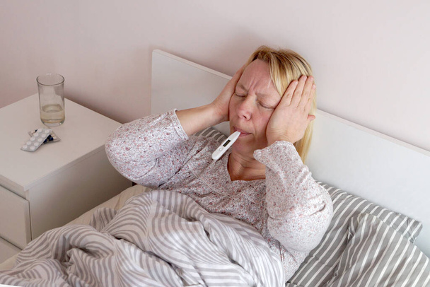 Μια άρρωστη γυναίκα μετράει τη θερμοκρασία κρατώντας τα χέρια της στο πονεμένο κεφάλι της.. - Φωτογραφία, εικόνα