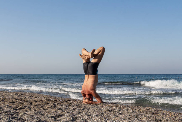 Middle age Man in shorts is doing his morning exercises on the beach by the sea. Headstand - Photo, Image