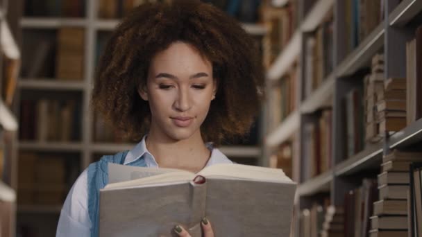 Молода афроамериканська студентка стоїть у публічній бібліотеці університету, зацікавлена в читанні книжки цікавої історії, уважно дивлячись на підручник, перегортаючи сторінку, щоб вивчати літературу, яка любить читати - Кадри, відео