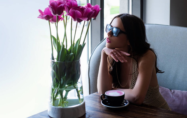 Romantic woman with coffee cup. Young stylish woman, fashion sunglasses, sitting in cafe with cup cappuccino. Spring tulips. - Photo, Image