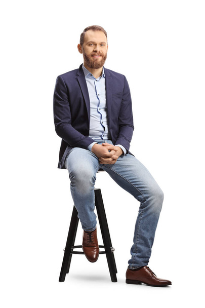 Full length portrait of a man in a suit and jeans sitting on a bar chair isolated on white background - Фото, зображення