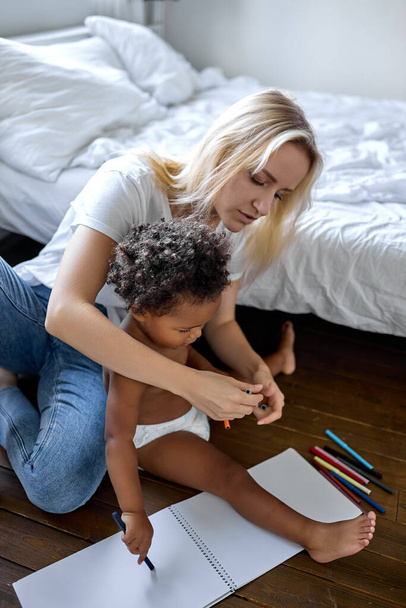 blonde female and small adorable daughter sitting on wooden warm floor drawing - Foto, Bild