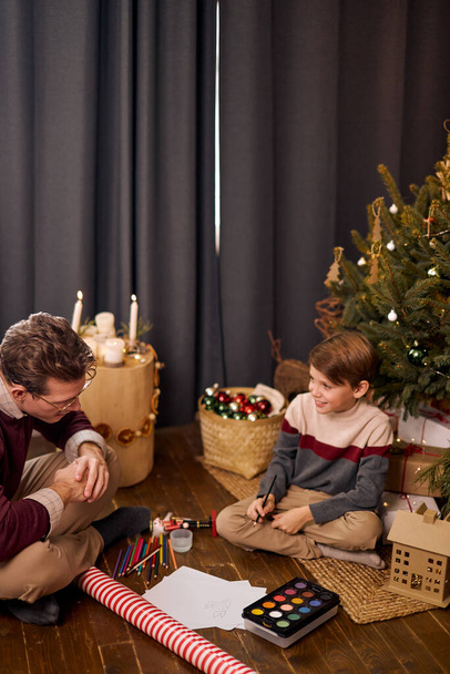 Nice Dad and Son preparing for christmas celebration. drawing on floor near xmas tree - Foto, imagen