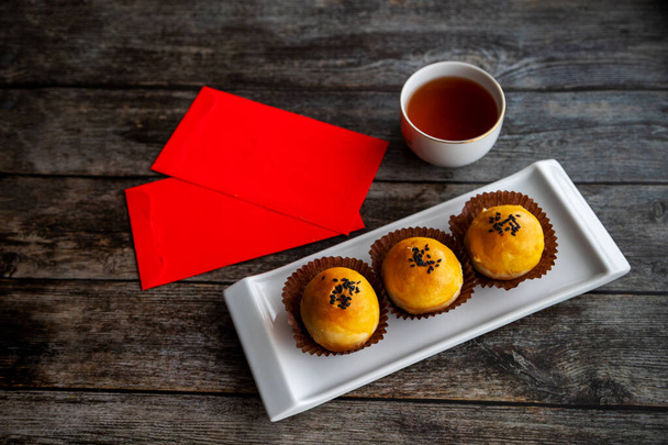 Tradiční čínské novoroční pečivo pečené s nadýchaným těstem a plněné slaným žloutkem a sladkou pastou. Čínské štěstí peníze červené balíčky a čaj v pozadí na rustikální dřevěný stůl. - Fotografie, Obrázek