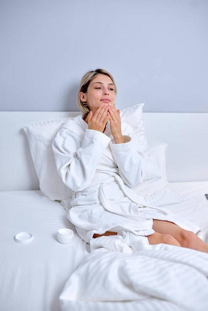 young lady in bathrobe applying cream on face touching moisturized healthy sensitive skin - Fotografie, Obrázek