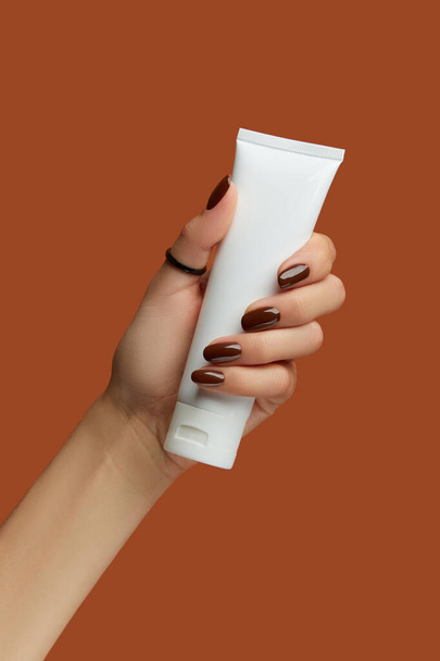 Womans hand holding white tube on brown background. Self care concept. - Fotó, kép