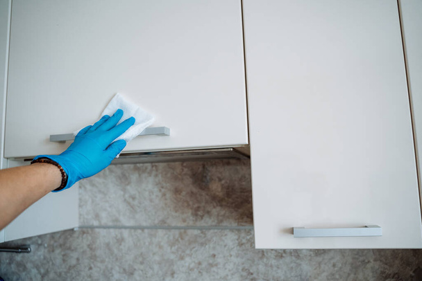 Cleaning of cabinets in the kitchen with a damp cloth. Wipe door handles with detergent. Disinfection of surfaces. Hand with rag. Blue glove. High quality photo - Photo, Image