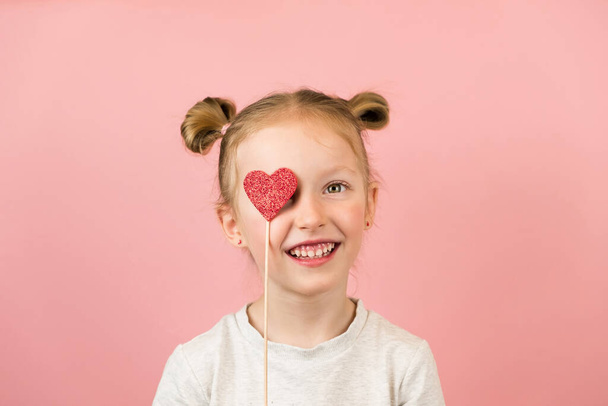 Funny little blonde girl smiling and playing with red heart toy on pink background. Valentines Day or Mothers Day concept - Foto, Imagen