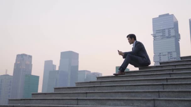 ázsiai üzletember ül a lépcsőn a mobiltelefon és gondolkodás modern épület háttér - Felvétel, videó