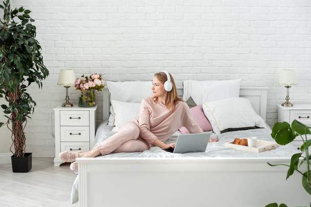 Pleasant middle-aged blonde woman in a home suit and headphones sitting in bed with a laptop. Work and study from home. Online communication. Distance education. Space for text. - Foto, Imagem