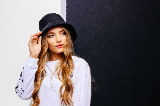 A beautiful young girl looks with interest and holds a hat with her hand. A model with long hair and blue eyes in a hat against a black and white wall with copy space in the middle. - Φωτογραφία, εικόνα