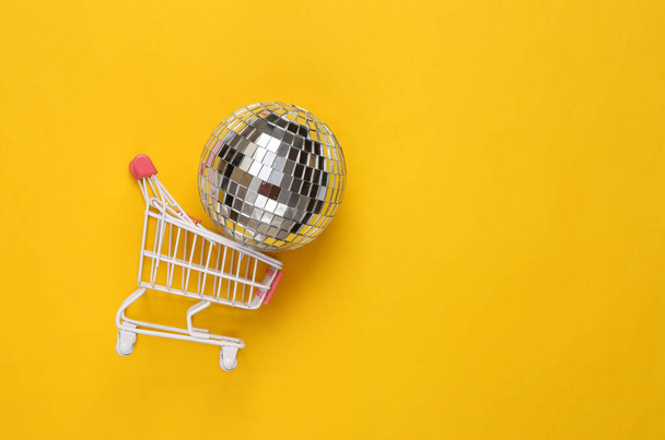 Shopping cart with disco ball on yellow background. Party concept. Creative minimal layout. Top view - Fotografie, Obrázek